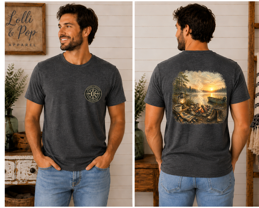 Man wearing the Campfire Sunrise graphic tee featuring a peaceful lakeside campfire scene with boots, a blanket, lantern, coffee cup, and sunrise.